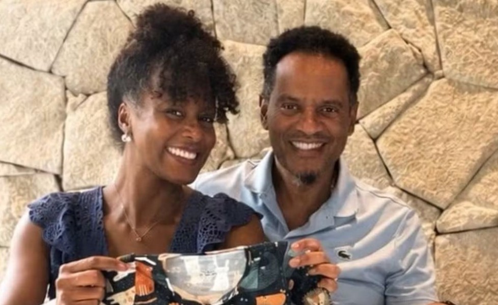 Maju Coutinho, jornalista da Globo, ao lado do marido, segurando uma roupinha de bebê, anuncia que está grávida.