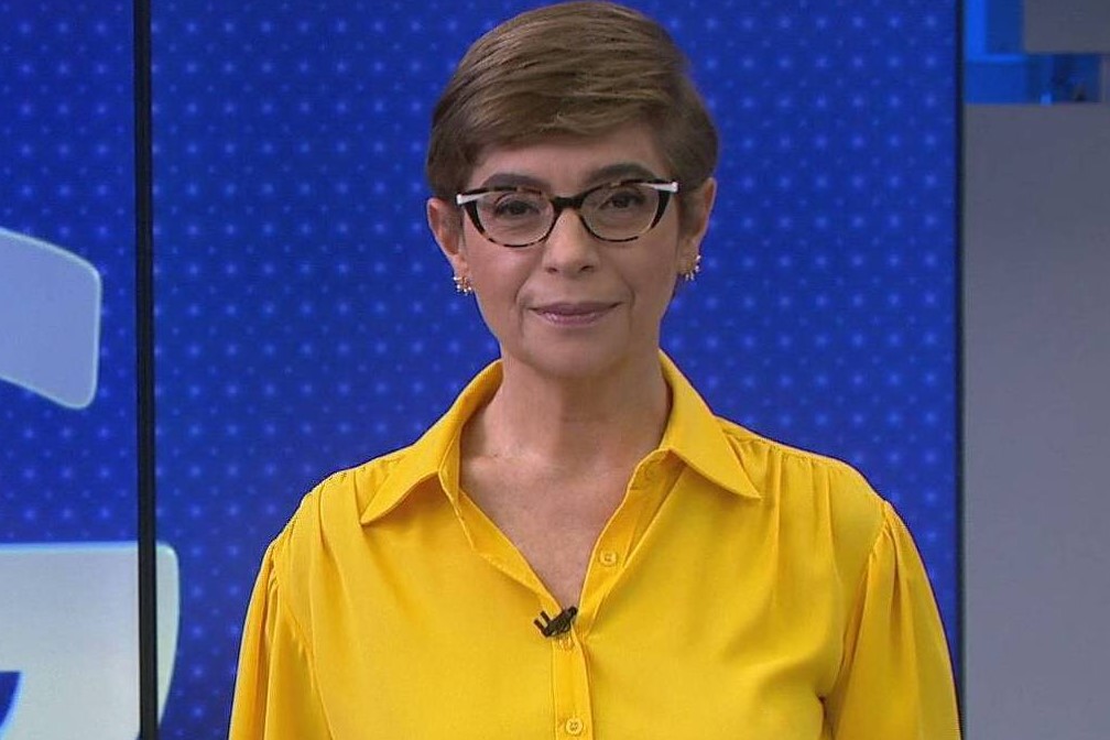 Âncora do Jornal da Globo, Renata Lo Prete posa no estúdio do jornalístico, usando uma blusa da cor amarela e com um tímido sorriso.